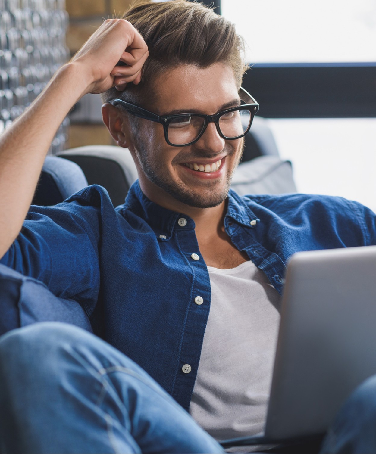 Ein Mann sitzt auf einem Sofa, lächelt und schaut auf etwas auf einem Laptop.